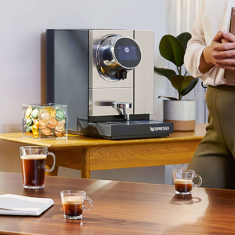 A sleek silver and black Nespresso Momento 100 Commercial Coffee Machine featuring capsule recognition is placed on a wooden counter. There are clear glass mugs of coffee on the counter and table. A person holding a tablet is standing near the machine, and a clear container with colorful coffee capsules is also visible.