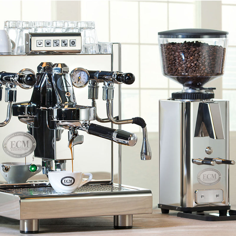 An ECM Mechanika Max Heat Exchanger (HX) Espresso Machine with PID is brewing coffee into a white cup, beside an ECM S-Automatik 64 Burr Grinder filled with beans. Both are set on a wooden countertop, illuminated by bright window light in the background.