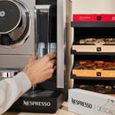 A person is using a Nespresso Professional Momento 100 Commercial Coffee Machine by Nespresso. They are placing a cup in the dispenser. To the right, there is a rack with various Nespresso coffee pods in colorful boxes. The surroundings are part of a modern kitchen or coffee station, featuring advanced touchless technology.