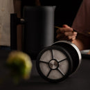 A close-up of a cylindrical coffee filter lying on its side on a dark surface, with the Fellow Clara French Press (Matte Black + Walnut) blurred in the background, exuding barista-level functionality. Partially visible people holding a spoon are seen as well. The foreground showcases a blurred green and yellow flower.