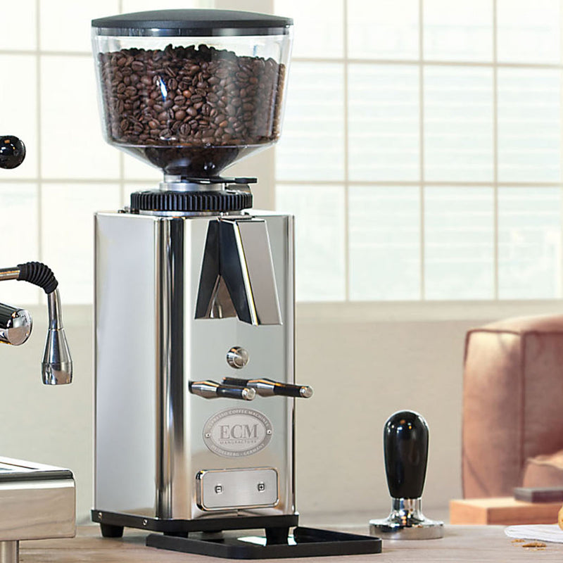 The ECM S-Automatik 64 Burr Grinder in sleek stainless steel rests on the countertop, filled with freshly ground coffee. Positioned next to a tamper and part of an espresso machine, this on-demand grinder by ECM is perfectly situated by a large window, ready to brew your perfect cup.