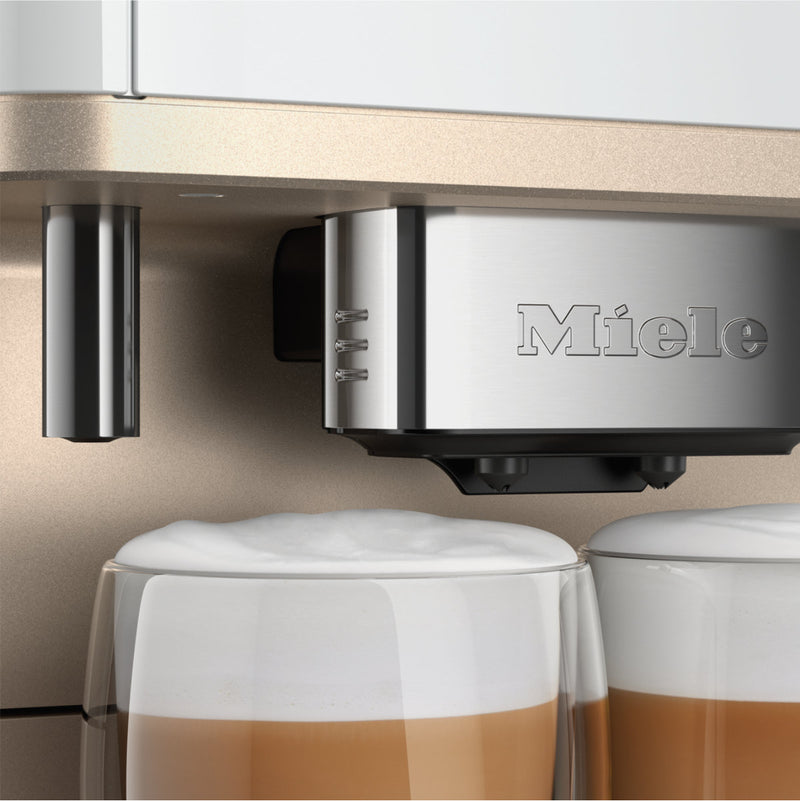 Close-up of a Miele CM6360 Super Automatic Countertop Coffee & Espresso Machine in Lotus white dispensing frothy milk into two clear glass mugs filled with layered coffee. The machine's elegant finish and the Miele logo are visible, and the mugs are tall and cylindrical with a rich, creamy top layer.