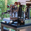 An ECM Mechanika VI Slim Espresso Machine - Demo, Unused, with its sleek stainless steel construction, multiple control knobs, and steam wand, sits on a dark counter. To the right of it stands a coffee grinder filled with beans, ready for brewing coffee and frothing milk. A window displaying lush greenery provides the perfect backdrop.