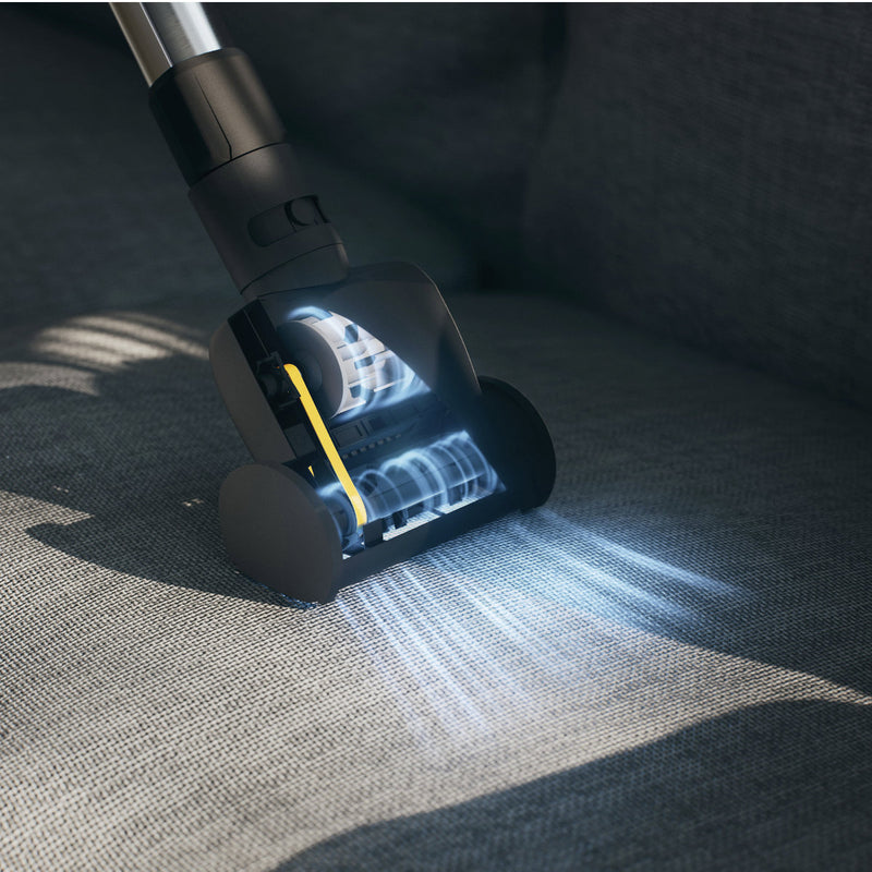 The Miele Handy TurboBrush Vacuum Cleaner Attachment STB 101, equipped with rotating brushes and LED lights, is used on the sofa to effectively clean the fabric surface. Sunlight casts soft shadows on the sofa, emphasizing its eco-friendly design.