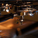 Close-up of a highly polished, stainless-steel portafilter and group head from the FAEMA Prestige Plus 2 Group Head Tall Cup Autosteam Espresso Machine F01-UQ210T2BTDDGA (Indigo Blue). The adjustable temperature control ensures perfect brewing. The background is softly illuminated with warm, golden lighting, creating a cozy and inviting atmosphere.