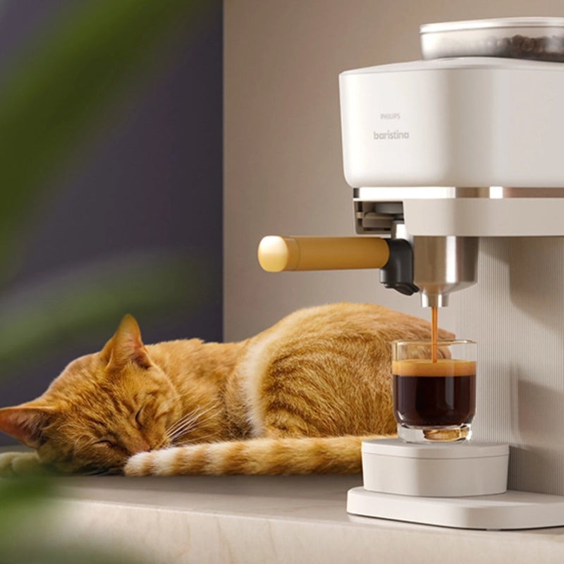 A ginger cat is sleeping on a countertop next to the Philips Baristina Combo Espresso Machine with Milk Frother BAR303/00 (White), which is in the process of dispensing barista-quality espresso into a glass cup. The scene, enhanced by the 16 bar pump pressure and soft lighting, feels peaceful and cozy with the aroma of fresh ground beans filling the air.