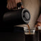 A person is seen pouring coffee from a sleek, matte black Fellow Clara French Press with walnut accents into a clear glass cup, showcasing barista-level functionality. The background is dark, and the focus is on the hand holding the French Press and the stream of coffee being poured. The person is wearing a light-colored shirt.