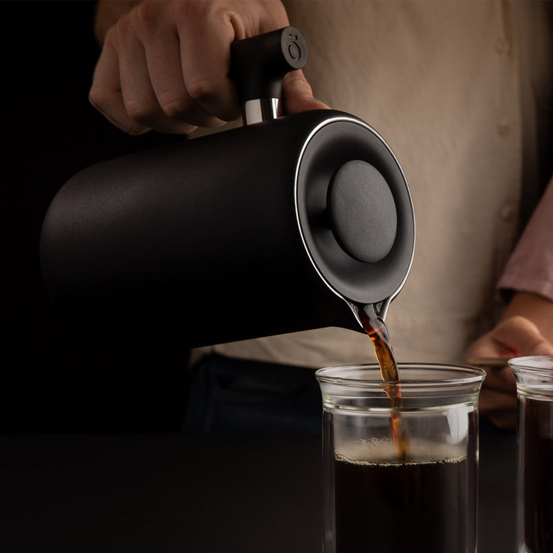 A person is seen pouring coffee from a sleek, matte black Fellow Clara French Press with walnut accents into a clear glass cup, showcasing barista-level functionality. The background is dark, and the focus is on the hand holding the French Press and the stream of coffee being poured. The person is wearing a light-colored shirt.