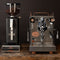 An ECM Mechanika VI Slim Espresso Machine from the ECM Heritage Line, showcasing an ECM-Customized E61 Group Head along with various dials and levers, is placed on a wooden surface beside a black coffee grinder filled with beans. Both exhibit a vintage design aesthetic.