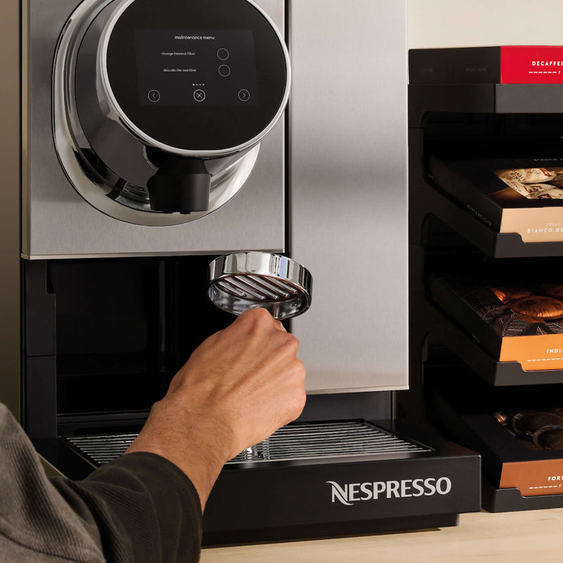 A person operating the Nespresso Momento 100 Commercial Coffee Machine is placing a portafilter underneath the coffee dispenser. The professional coffee machine, featuring a digital display and capsule recognition technology, stands to the right with stacks of Nespresso capsules clearly visible. The word "Nespresso" is prominently displayed on the base.