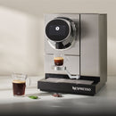 A Nespresso Momento 100 Commercial Coffee Machine effortlessly brews a shot of espresso into a clear glass cup using its touchless technology. Nearby, another clear glass mug holds freshly brewed coffee, while two Nespresso coffee pods—one green and one brown—rest on the counter. The sleek, modern machine boasts a professional stainless steel design.