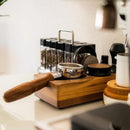 A wooden portafilter stand holds a portafilter and the Normcore Narrow Espresso Tamping Station - Walnut. In the background, a set of spice jars with black lids sits on the counter next to a coffee grinder and other 58mm and 54mm espresso accessories. The countertop is clean and organized.