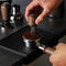 A person uses the Breville Distribution Duo™ 54mm BEA203WLW to evenly spread coffee grounds in a portafilter on a dark surface. Nearby, coffee tampers and a Temp Control Milk Jug with a latte art spout elevate the barista's setup, ensuring perfectly crafted drinks every time.