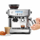 A Breville The Barista Pro Espresso Machine BES878 in Olive Tapenade, featuring a stainless steel design and a digital display, is brewing espresso into a glass. Nearby, hands are frothing milk in a stainless steel cup using its ThermoJet heating system. A cup of coffee and a pastry are placed beside the semi-automatic machine.