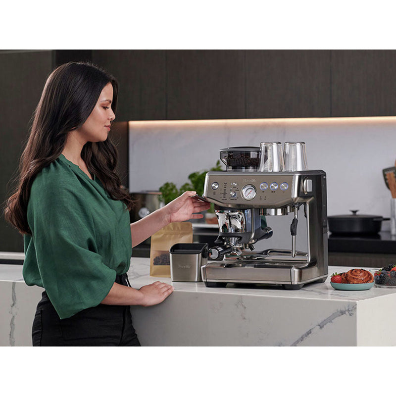 A woman in a green blouse operates the Breville Barista Express Impress Semi-Automatic Espresso Machine BES876OTL (Olive Tapenade) on a white marble countertop in a modern kitchen. Next to her are coffee beans in a brown bag and two pastries on a small plate. Another kitchen counter with various items is visible in the background.