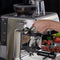 A person operating a Breville The Barista Express Impress Semi-Automatic Espresso Machine BES876OTL (Olive Tapenade) places a portafilter beneath the coffee grinder. The olive tapenade machine features various buttons and settings. Fresh coffee beans are visible in the bean hopper on top, with fresh fruits in the background.