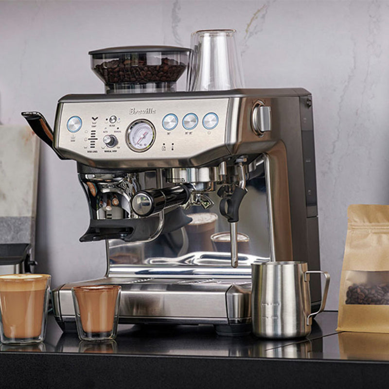 The Breville The Barista Express Impress Semi-Automatic Espresso Machine BES876OTL in Olive Tapenade is placed on a kitchen countertop. Two cappuccinos and a metal milk frothing pitcher are in front of it, showcasing its automatic dosing feature for perfect brews. A beige coffee bag is situated to the right of the machine.