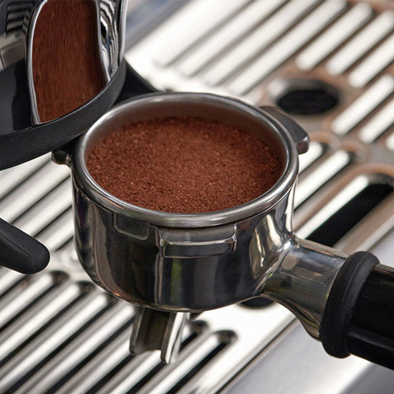 Close-up of a Breville The Barista Express Impress Semi-Automatic Espresso Machine BES876OTL (Olive Tapenade) portafilter containing freshly tamped coffee grounds, ready for brewing. Utilizing Breville's assisted tamping technology, the portafilter is positioned against a metallic surface with a grill-like design in the background.