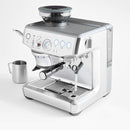 The Breville The Barista Express Impress Semi-Automatic Espresso Machine BES876OTL in Olive Tapenade features a portafilter, frothing wand, and bean grinder on top. Several control buttons and a pressure gauge are visible on the front, along with automatic dosing capabilities. A stainless steel milk frothing pitcher is placed beside the machine on a white surface.