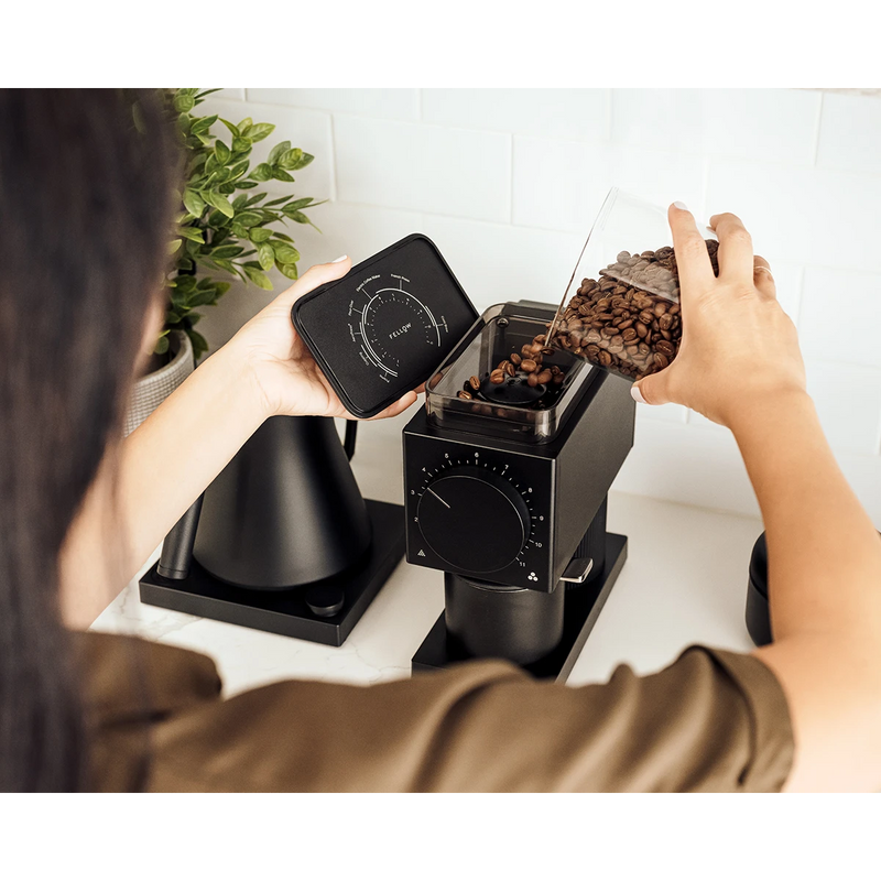 A person is pouring coffee beans from a container into a Fellow Ode Coffee Grinder Gen 2.0 (Matte Black) on a kitchen counter. The person holds the lid of the grinder in their other hand. The scene is set against a white tiled backsplash with a potted plant and other kitchen appliances nearby, setting the stage for perfect brewed coffee.