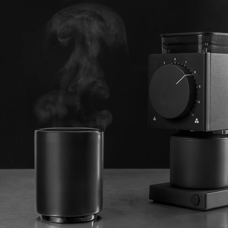 A Fellow Ode Coffee Grinder Gen 2.0 (Matte Black) is elegantly placed on a counter beside a steaming black mug, set against a dark background. The scene evokes the preparation of freshly ground coffee, with adjustable grind settings for the perfect cup courtesy of Fellow.