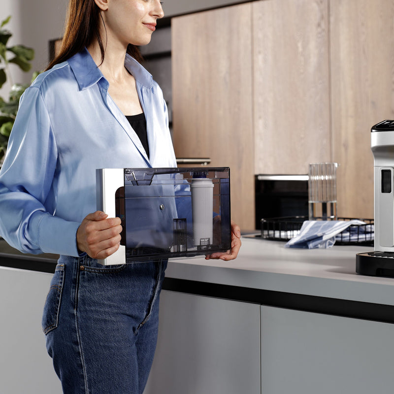 A person wearing a light blue shirt and jeans is holding a DeLonghi Water Filter for ECAM Type Espresso Machines (DLSC002) - 3 Pack in a modern kitchen setting. The filter, known for reducing limescale buildup and improving machine efficiency, is held next to a countertop with various kitchen appliances and utensils visible in the background.