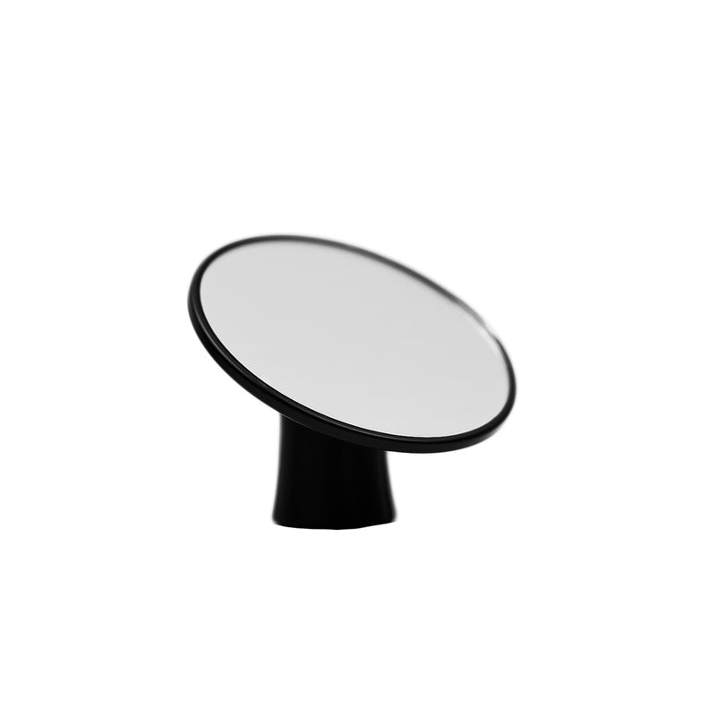 The HCS Espresso Shot Mirror, designed for bottomless portafilters, features a circular, reflective surface mounted on a black base and stands prominently against a white background. Reminiscent of an espresso shot being poured, the mirror is slightly tilted upward and to the side.