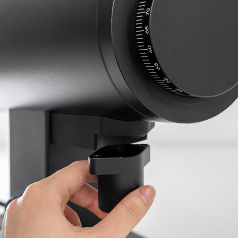 A close-up shows a person adjusting the DF Grinders DF83V, a black coffee grinder with DLC brew burrs, by turning the dial. It boasts a sleek design with visible adjustment scale and variable speed control. The background is blurred.