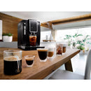 A modern black DeLonghi Dinamica Super Automatic Espresso & Coffee Machine - ECAM35020B (Black) - Open Box, Unused is placed on a wooden table in a bright, airy kitchen. It is dispensing iced coffee into a glass. Various types of coffee drinks, including espresso, cappuccino, and iced coffee, are arranged in clear glass cups along the table.