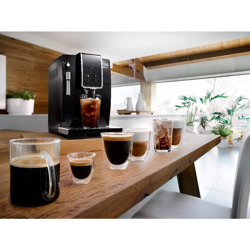 A modern black DeLonghi Dinamica Super Automatic Espresso & Coffee Machine - ECAM35020B (Black) - Open Box, Unused is placed on a wooden table in a bright, airy kitchen. It is dispensing iced coffee into a glass. Various types of coffee drinks, including espresso, cappuccino, and iced coffee, are arranged in clear glass cups along the table.