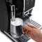 A person's hand holding a stainless steel frothing pitcher filled with frothy milk under the steaming wand of a black DeLonghi Dinamica Super Automatic Espresso & Coffee Machine - ECAM35020B. The machine's control panel and drip tray are visible in the background, showcasing advanced De'Longhi Brewing Technology.