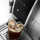 A close-up of the DeLonghi Dinamica With Adjustable Frothing Wand Super Automatic Espresso & Coffee Machine ECAM35025SB (Silver) - Open Box, Unused, pouring iced coffee into a transparent glass filled with ice cubes. The DeLonghi machine features a control panel with several buttons and is mounted on a sleek metal drip tray. The coffee appears dark and rich.