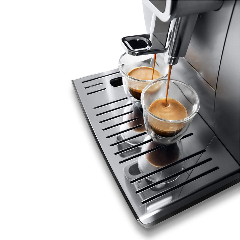 A close-up of an open-box, unused DeLonghi Dinamica With Adjustable Frothing Wand Super Automatic Espresso & Coffee Machine ECAM35025SB (Silver) brewing two cups of coffee simultaneously. The espresso is being dispensed into two glass cups placed on a shiny metallic drip tray, with a perfect layer of crema visible on both coffees.