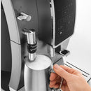 A close-up view of a super automatic coffee machine frothing milk into a stainless steel pitcher, held by a hand. The DeLonghi Dinamica With Adjustable Frothing Wand Super Automatic Espresso & Coffee Machine ECAM35025SB (Silver) - Open Box, Unused features a digital touch screen interface and a sleek, metallic design. The text "HOT MILK CAPPUCCINO" is visible on the frothing nozzle.