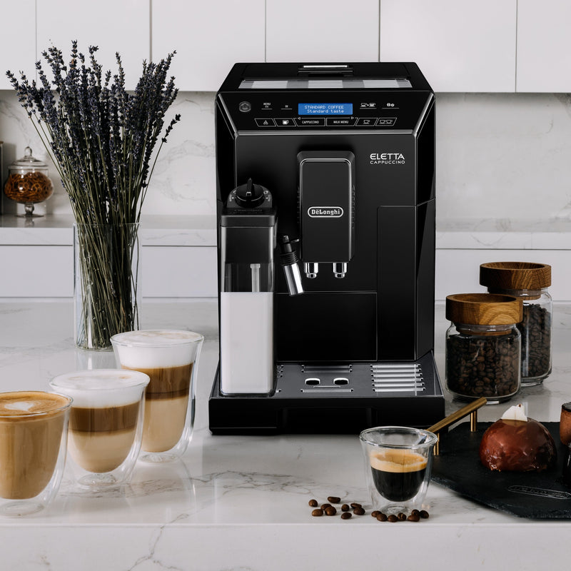 A refurbished DeLonghi Eletta Cappuccino Super Automatic Espresso Machine ECAM44660B (Black) sits on a white marble countertop, complete with a milk frother. Four glass mugs filled with barista-quality drinks are arranged around it, accompanied by jars of coffee beans, a small glass of espresso, a red dessert, and a vase of lavender.