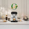 A modern kitchen countertop showcases a DeLonghi Eletta Explore Super Automatic Espresso Machine with Cold Brew ECAM45086S expertly crafting a cappuccino. The space is adorned with vases, a sealed jar, cookies, and various coffee drinks. Above it hangs a wreath featuring a ribbon that reads "Consumer Favorite Coffee 2023," honoring its mastery in super automatic espresso brewing.