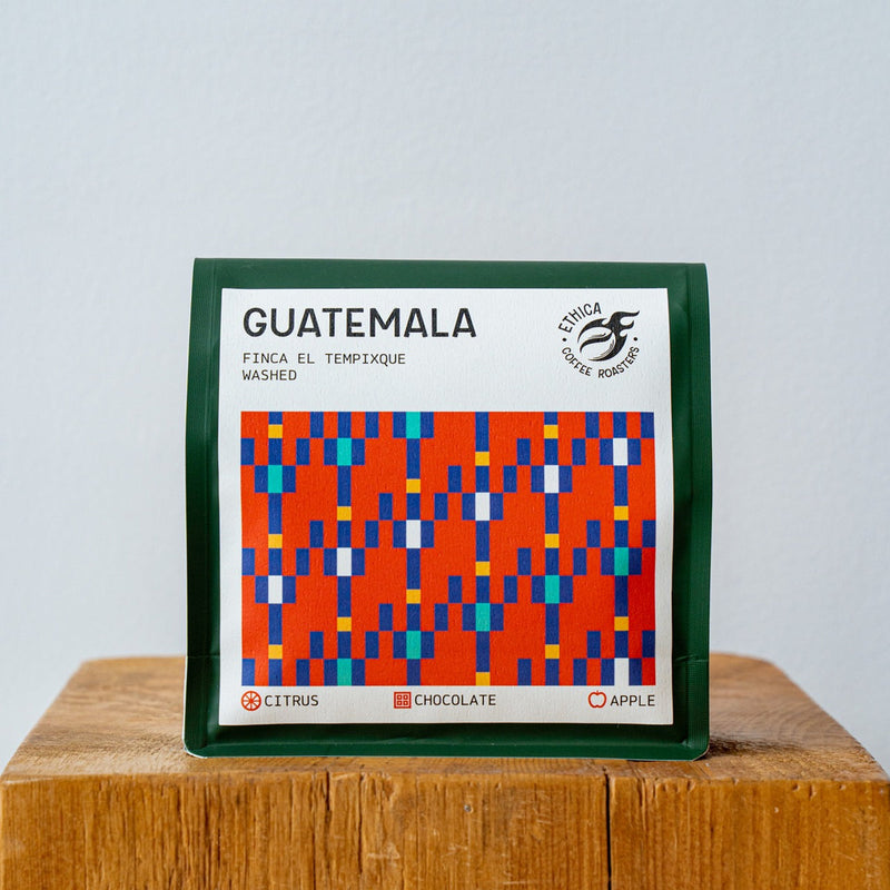 The 250g bag of Ethica Coffee Roasters Guatemala Finca El Tempixque Washed Whole Coffee Beans, adorned with vibrant geometric patterns in red, blue, and orange, highlights distinctive flavor notes of citrus, chocolate, and apple. The bag is positioned on a wooden surface against a plain background.