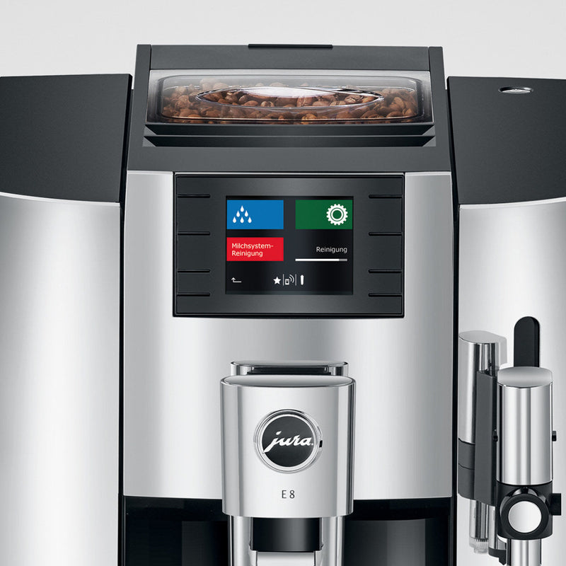 A sleek chrome Jura E8 Chrome Automatic Coffee Machine 15371 OPEN BOX (4033) is shown from the front. The LCD screen displays maintenance options with colored icons, including settings for the Intelligent Water System (I.W.S.). The top compartment is filled with coffee beans, and the Jura logo is prominently displayed in the center.