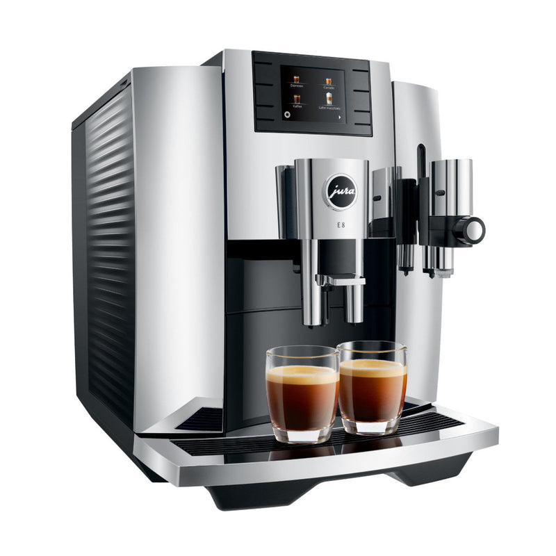 A sleek, open-box Jura E8 Chrome Automatic Coffee Machine brews two cups of espresso. Featuring a digital display and buttons for various brewing options, it includes the Pulse Extraction Process (P.E.P.) for optimal flavor and a milk frother on the right side. Two glass cups filled with espresso sit on its tray.