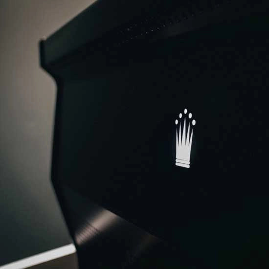 A close-up of the Conti MC Ultima Espresso Machine (Black) showcases a minimalist white crown-shaped logo at the center, exuding elegance and luxury. The dark, blurred background enhances the refined charm of this high-end espresso machine's design.