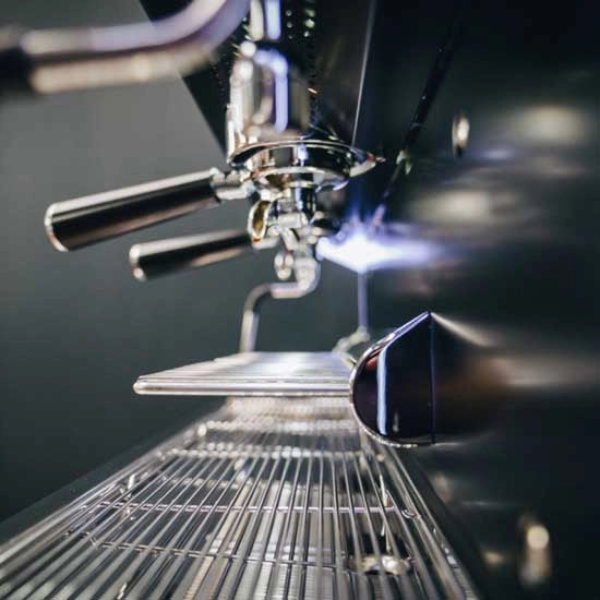 Close-up of the Conti MC Ultima Espresso Machine (Black), featuring sleek, modern design elements like metallic levers and a grill tray, beautifully illuminated by soft lighting. The angle emphasizes its intricate craftsmanship, including pre-infusion capabilities and adjustable temperature controls for precise coffee-making.