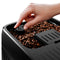 A hand adjusts the grind size selector knob on a sleek, modern DeLonghi Magnifica Plus Super Automatic Espresso Machine with LatteCrema System (ECAM32070SB), which is filled with beans. The glossy black machine, surrounded by coffee beans, boasts 18 coffee drinks and advanced LatteCrema Hot technology for the perfect cup every time.