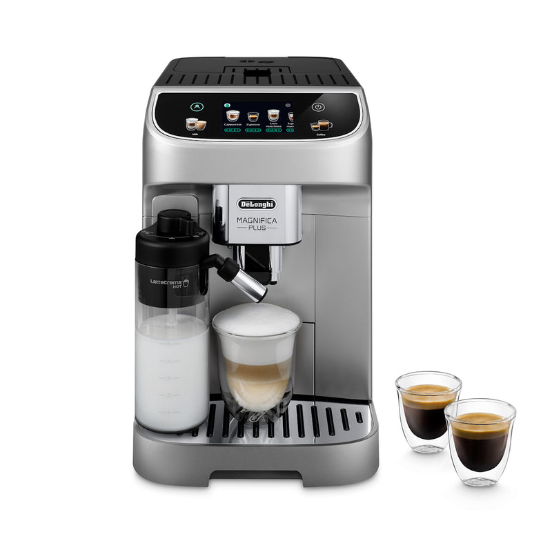 A DeLonghi Magnifica Plus Super Automatic Espresso Machine with LatteCrema System (ECAM32070SB) - PREORDER is positioned on a flat surface. The machine, equipped with a milk frother attachment filled with milk, is brewing a latte into a glass mug. Two small glasses of espresso are placed to the right of the machine, demonstrating its capability to make 18 different coffee drinks.