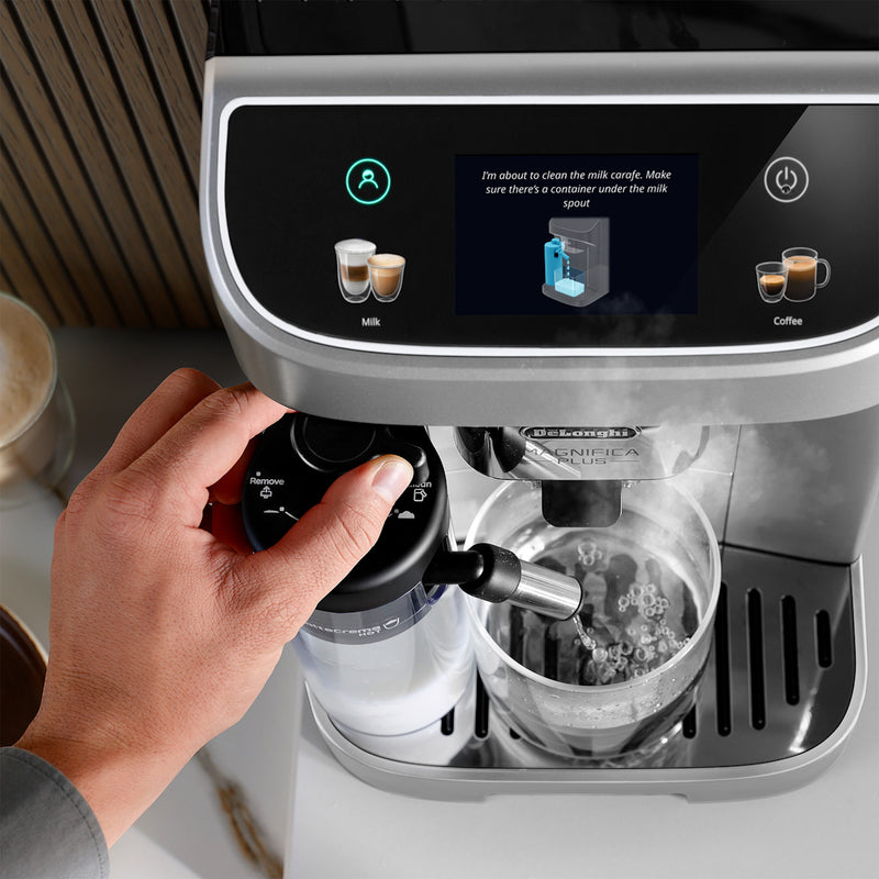 A person is using the DeLonghi Magnifica Plus Super Automatic Espresso Machine with LatteCrema System (ECAM32070SB) from DeLonghi, which features a touchscreen display displaying cleaning instructions for the milk carafe. The person's hand is holding the milk frother knob on the left side of the machine, utilizing LatteCrema Hot technology while a cup is placed under the frother.