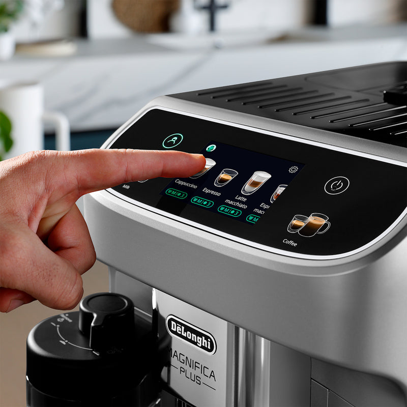 Close-up of a person’s hand selecting a coffee option on the touch screen of a DeLonghi Magnifica Plus Super Automatic Espresso Machine with LatteCrema System (ECAM32070SB). Various choices like cappuccino, espresso, and one of the 18 available coffee drinks are displayed. A coffee cup is positioned below the machine's spout.