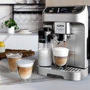 A DeLonghi Magnifica Plus Super Automatic Espresso Machine with LatteCrema System (ECAM32070SB) dispenses coffee into a cup with frothed milk on a kitchen counter. Two glass mugs with layered coffee and foam sit nearby. The background features a plant, a fruit bowl, and a shelf with glassware.