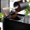 A hand pours coffee beans from a black canister into the bean hopper of a DeLonghi Magnifica Plus Super Automatic Espresso Machine with LatteCrema System (ECAM32070SB). The background features a wicker chair, a window bathed in natural light, and green potted plants, creating a cozy ambiance ideal for enjoying one of 18 coffee drinks.