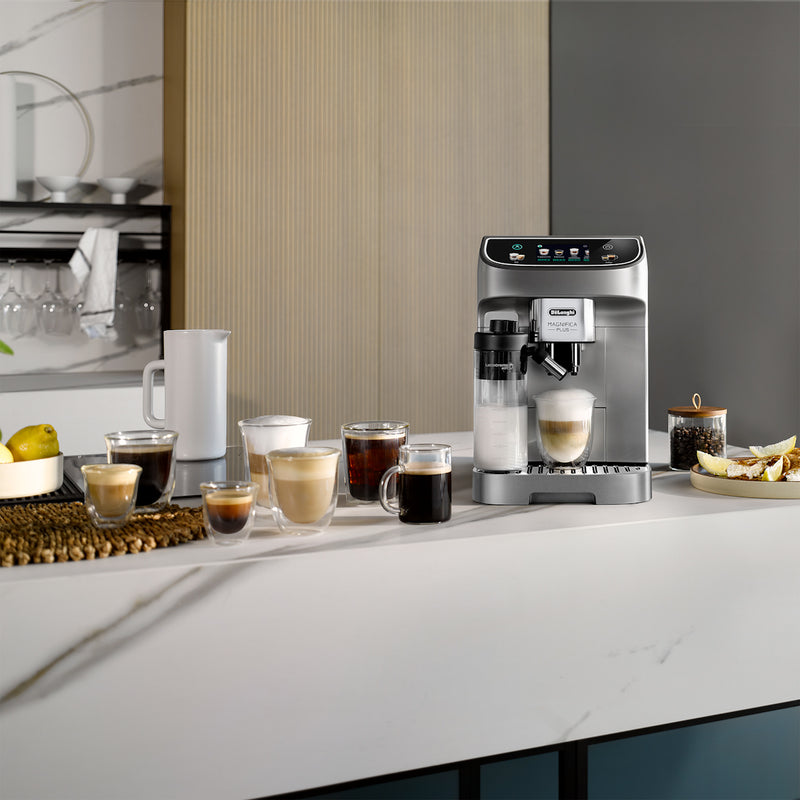 A modern kitchen countertop showcasing the DeLonghi Magnifica Plus Super Automatic Espresso Machine with LatteCrema System (ECAM32070SB) - PREORDER, seamlessly crafting a latte. Various coffee drinks, including cappuccino, espresso, and black coffee, are placed alongside a pitcher and a plate of food. Shelving in the background holds dishes and decor.