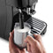 A person uses the DeLonghi Magnifica Start Super Automatic Espresso Machine ECAM22022B (Black) to effortlessly craft creamy coffee drinks, holding a stainless steel milk frothing pitcher under the steam wand. The pitcher is partially filled with frothed milk, highlighting the LatteCrema Hot perfection, while integrated grinding technology ensures precision brewing.