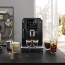 A DeLonghi Magnifica Start Super Automatic Espresso Machine ECAM22022B in black rests on a kitchen counter, utilizing its integrated grinding technology to brew espresso into two small glasses. To the right, glasses filled with espresso and iced coffee highlight the machine's versatility. The backdrop reveals a modern, sunlit kitchen extending gracefully.